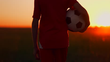 Young-boy-football-player-in-a-red-t-shirt-is-with-a-soccer-ball-on-the-field-at-sunset